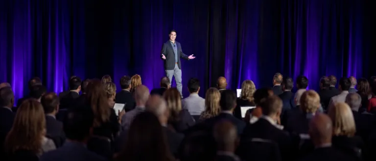 Paul Takisaki on stage in front of a deep-purple curtain, addressing a seated executive audience. Wearing a charcoal blazer over a dark patterned shirt, open-handed mid-gesture. Founder, former Verizon AVP.